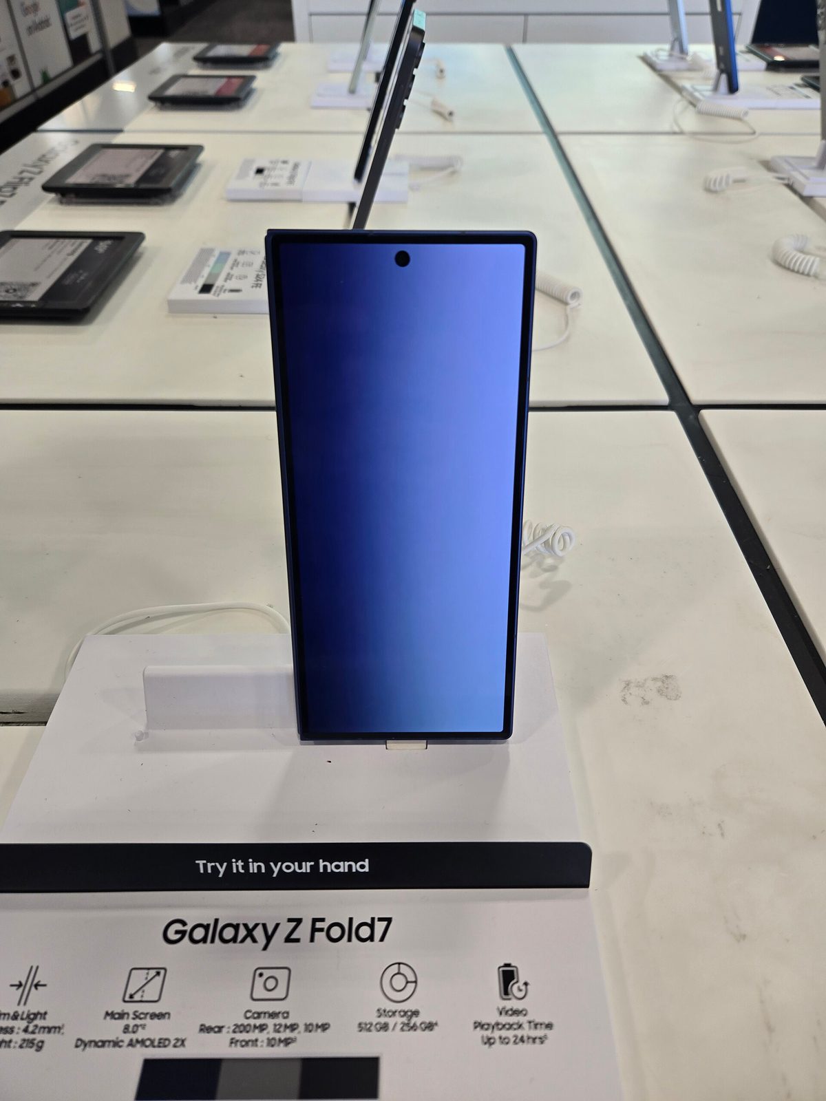 Samsung Galaxy Z Fold7 on a retail display stand, with its cover screen showing and the "Galaxy Z Fold7" signage and spec sheet visible below it